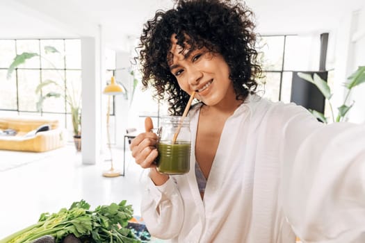Young african american woman taking selfie drinking green juice reusable bamboo straw in bright loft apartment. Home concept. Technology concept. Health concept. Copy space