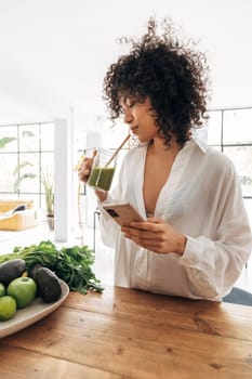 Young african american woman looking at her phone, browsing through social media while having a green juice. Reusable bamboo straw.Vertical. Home concept. Technology concept.