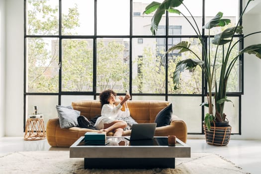 Young African American woman sitting on a couch holding cellphone waving hand video calling distance friend. Home concept. Technology concept. Copy space