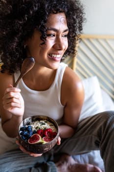 Vertical portrait of happy young latina woman smiling eating healthy breakfast bowl of oats and fruit sitting on bed. Vertical image. Healthy lifestyle concept.