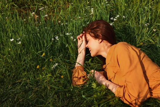 a calm woman with long red hair lies in a green field with yellow flowers, in an orange dress, smiling pleasantly, closing her eyes from the bright summer sun, resting her head on her hands. High quality photo