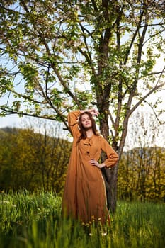 a sweet, attractive woman with long red hair stands in the countryside near a flowering tree in a long orange dress and looks at the camera holding her hand over her head. High quality photo