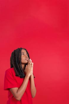 African woman looking up while praying with folded hands in studio with red background