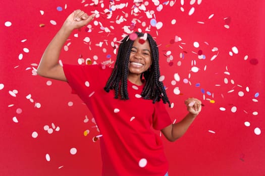 Happy african woman surrounded by confetti flying in the air in studio with red background