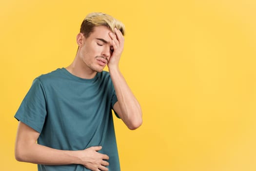 Gay man gesturing bodily discomfort and headache in studio with yellow background