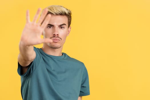 Gay man gesturing prohibition and stop with hand in studio with yellow background