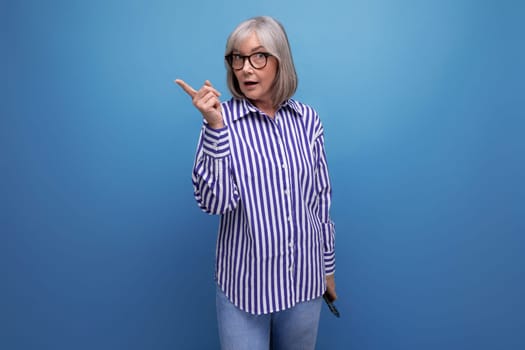 surprised middle aged woman with gray hair points her finger to the side on a bright studio background with copy space.