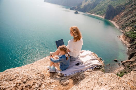 Freelance woman with her daughter working on a laptop by the sea, typing on the keyboard, enjoying the beautiful view, highlighting the idea of remote work