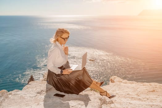 Business woman on nature in white shirt and black skirt. She works with an iPad in the open air with a beautiful view of the sea. The concept of remote work