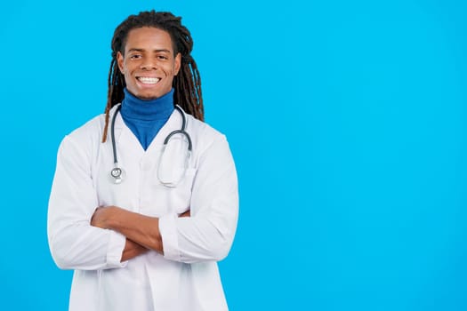 Latin male doctor with dreadlocks smiling at the camera with arms crossed in studio with blue background