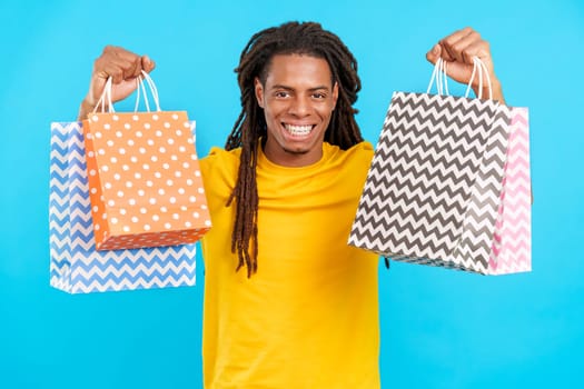 Latin man with dreadlocks raising shopping bags while smiling at camera in studio with blue background
