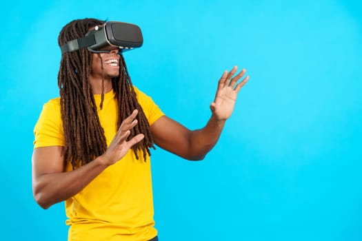 Happy latin man with dreadlocks wearing Virtual Reality goggles in studio with blue background