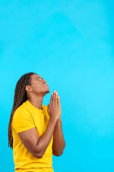 Latin man with dreadlocks looking up praying with folded hands in studio with blue background