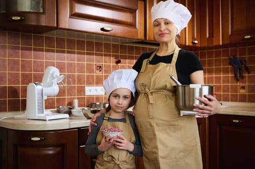 Happy pregnant woman, expectant mother and her lovely little daughter, dressed in apron and chef's hat, enjoying cooking together, standing in the home kitchen, smiling, confidently looking at camera