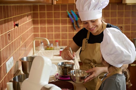 Happy smiling mother and daughter having fun together, preparing a homemade dessert, standing at kitchen counter, making delicious cake with melted belgian chocolate and whipped cream. Confectionery