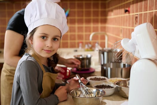 Caucasian little girl in chef's hat and kitchen apron, smiling, looking at camera, helping her mother to prepare a delicious homemade dessert for festive occasion. mom and daughter cooking together