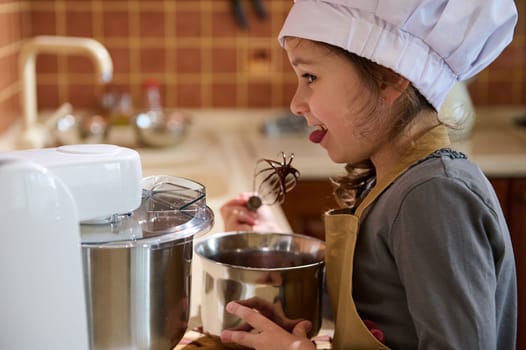Cute little girl helps her mom in kitchen to prepare cream for the cake, quietly licks a spoon with melted delicious confectionery chocolate, dressed as chef confectioner in white cap and beige apron