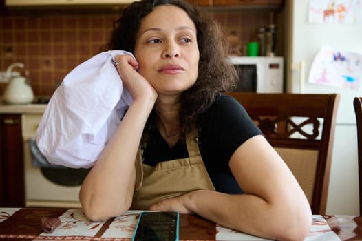 Beautiful pensive curly multi ethnic woman housewife, baker confectioner, chef pastry in chef's apron, sitting at table in home kitchen, cutely smiles looking away. Culinary. Cookery. People and hobby