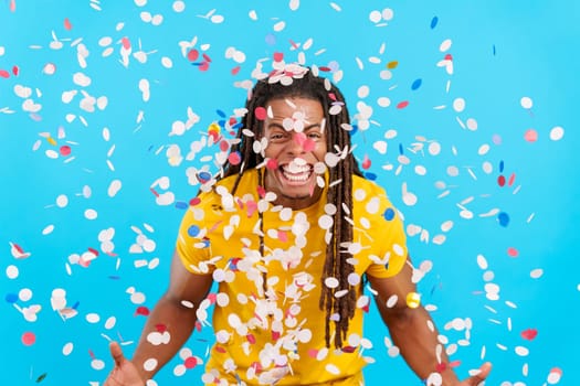 Happy latin man with dreadlocks surrounded by confetti in air in studio with blue background