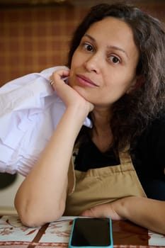 Close-up portrait of a multi ethnic young confident beautiful woman, baker confectioner in beige apron, holding a white chef's cap and looking at camera. Professional occupation. People.