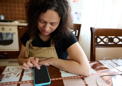 Pretty young woman wearing apron, amateur pastry chef, confectioner, housewife, sitting at kitchen table, checking mobile app with online recipe on her modern smartphone. People. Technology. Culinary