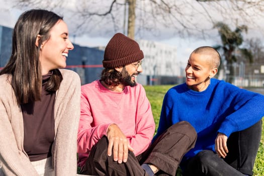 three young friends talking happy sitting in the lawn at the city park in a sunny day, concept of friendship and urban lifestyle