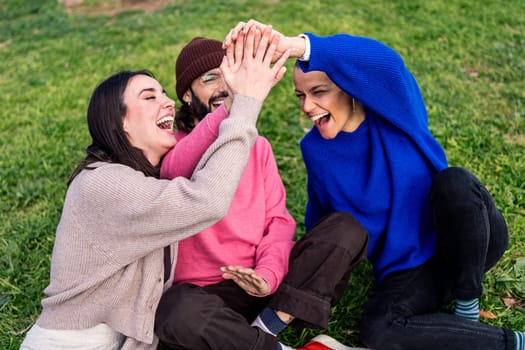 three young friends laughing funny high fives sitting on the grass, concept of friendship and modern lifestyle