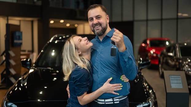 Happy caucasian couple hugging and man holding new car keys in car showroom