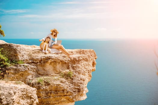 Freelance woman with her daughter working on a laptop by the sea, typing on the keyboard, enjoying the beautiful view, highlighting the idea of remote work