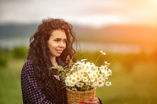 A woman stands on a green field and holds a basket with a large bouquet of daisies in her hands. In the background are mountains and a lake