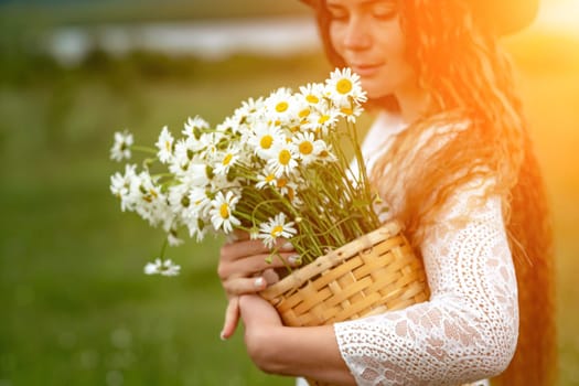 A middle-aged woman in a white dress and brown hat holds a lA middle-aged woman in a white dress and brown hat holds a basket in her hands with a large bouquet of daisies.arge bouquet of daisies in her hands. Wildflowers for congratulations.