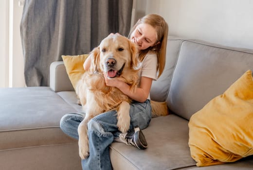 Preteen girl hugging golden retriever dog and smiling sitting on sofa at home. Beautiful child kid with labrador doggy pet indoors