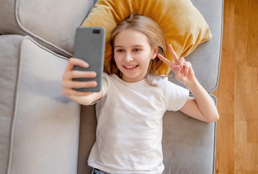Preteen girl with smartphone making selfie lying on sofa at home. Pretty child kid smiling with modern cellphone on couch