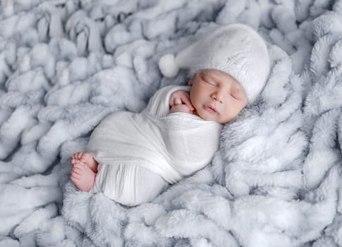 Cute newborn baby boy swaddled in grey fabric sleeping on fur blanket. Adorable infant child kid napping studio portrait