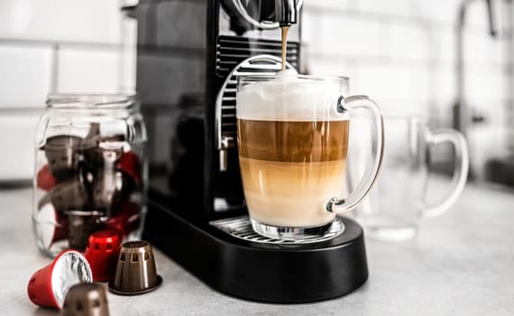 Coffee machine with capsules and creamy cappuccino in transparent cup at home. Espresso caffeine beverage maker and italian drink