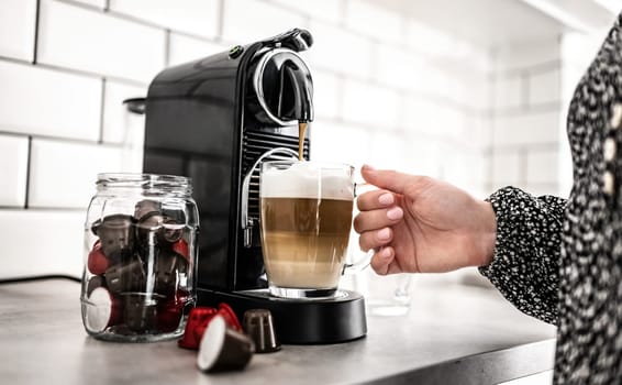 Girl with capsule coffee machine and cappuccino in transparent cup at kitchen. Woman preparing creamy italian caffeine beverage using professional espresso maker at home