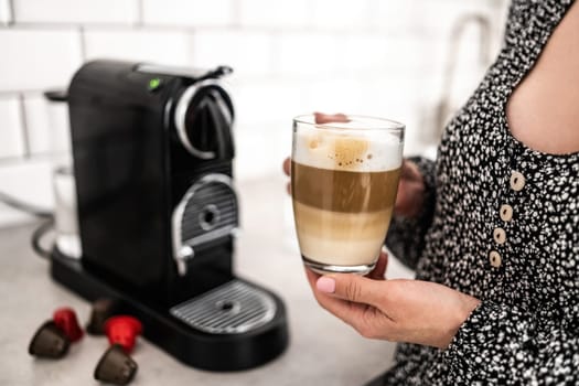 Girl with cappuccino in transparent cup and capsule coffee machine at kitchen. Woman holding mug with creamy italian caffeine beverage prepared in professional espresso maker