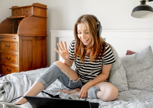 Pretty girl with headphones and laptop in bed during online learning webinar. Beautiful teenager talking by video call with pc computer at bedroom