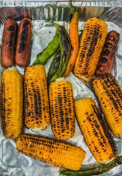 Top view of grilled corn cobs, green chili pepper and sausages on disposable aluminum  food packaging.