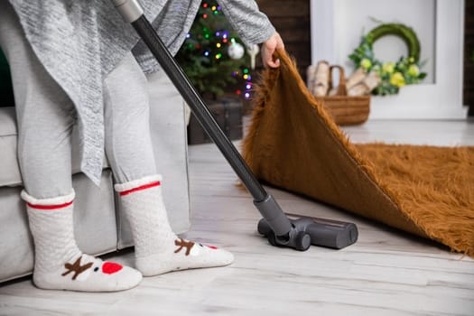 A woman lifts a rug up by the edge and vacuums thoroughly under it. The apartment decked out in decorations for reliving Christmas.