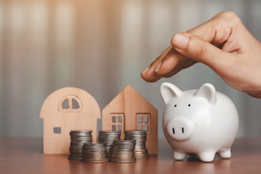 Hand covering a piggy bank with the wooden house model and a pile of coins and money on the table for business, finance, saving money and property investment concept.