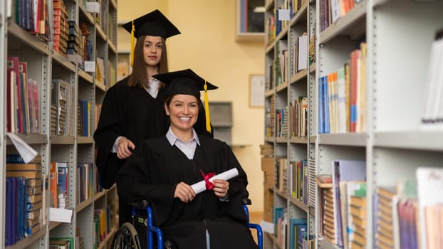 Happy young woman and woman in wheelchair in graduate gown with diploma in hands in library. Inclusive education