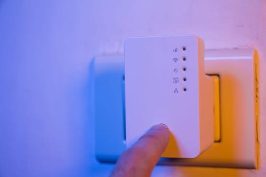 Man press with his finger on WPS button on WiFi repeater which is in electrical socket on the wall. The device help to extend wireless network in home or office.