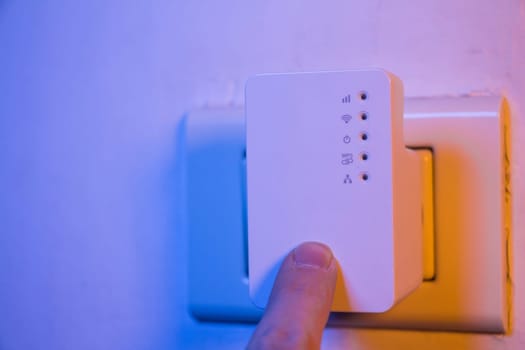 Man press with his finger on WPS button on WiFi repeater which is in electrical socket on the wall. The device help to extend wireless network in home or office.