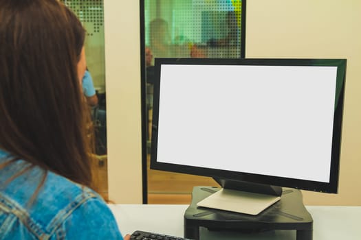 Close up back view of a young businesswoman looking at a screen while working on a desktop computer at her desk at the office. Modern and comfortable interior office. Mockup blank white screen with copy space.