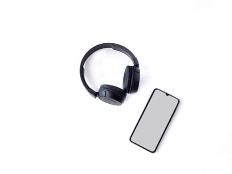 Black wireless headphone and mobile smartphone with a blank screen mockup lay on the surface of a white background. Top view flat lay with copy space. Music concept.