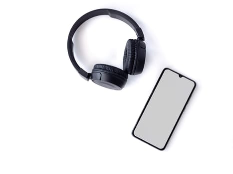 Black wireless headphone and mobile smartphone with a blank screen mockup lay on the surface of a white background. Top view flat lay with copy space. Music concept.