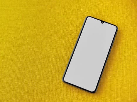 Black mobile smartphone mockup lies on the surface with a blank screen isolated on a yellow fabric background. Top view close up with selective focus and copy space.