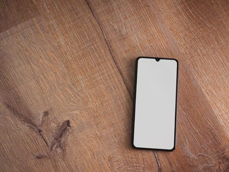 Black mobile smartphone mockup lies on the surface with blank screen isolated on wooden background. Top view flat lay with copy space.