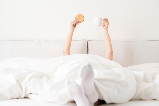 Female hand holding cup of coffee from under a blanket in bed. Woman waking up in the morning.